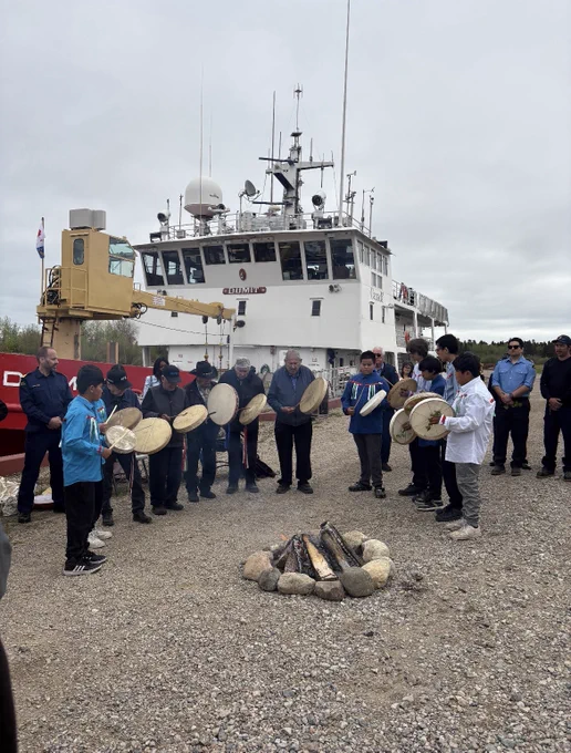 A group of people stands on a gravel shoreline in a semicircle around a small fire surrounded by stones, with light smoke rising. Several individuals hold round hand drums. In the background, a large red and white vessel with antennas and radar equipment is docked. The sky is overcast, and the setting appears to be a calm riverside or dock area.