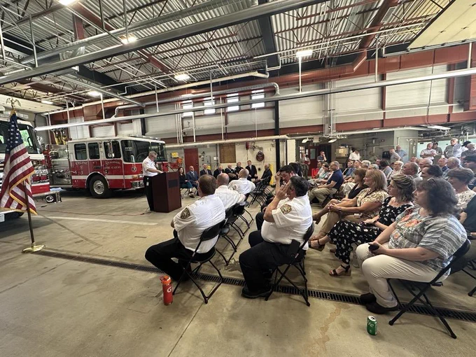 Wide angle shot of the inside of the NH Fire Academy Firehouse, with two firetrucks in the background, an American flag, and the Director of the Academy speaking at the podium to a gathered crowd of seated guests.