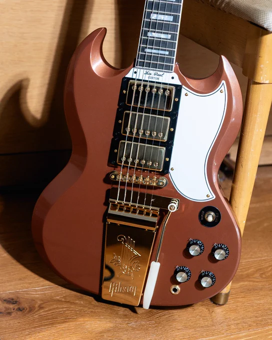 A Gibson SG electric guitar with a reddish-brown body is leaning against a chair. It features a white pickguard and a gold-colored tailpiece with the Gibson logo. Several control knobs are visible on the lower part of the body.