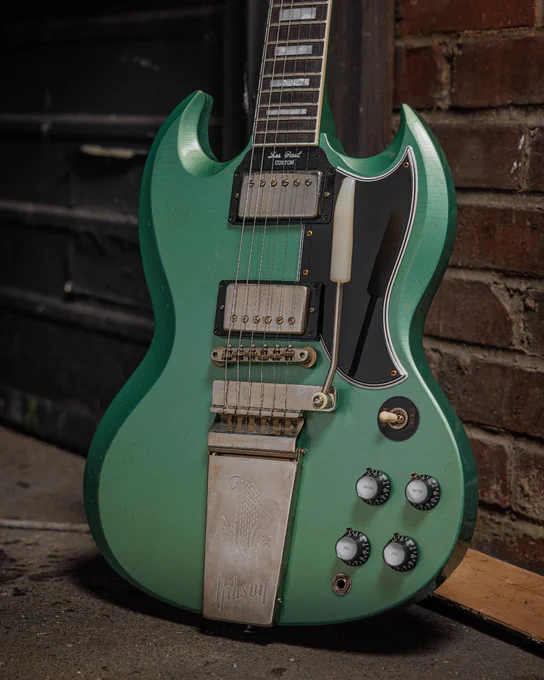 A green Gibson SG electric guitar leaning against a brick wall and metal door. The guitar has a distinctive double cutaway body, multiple control knobs, and a silver tailpiece.