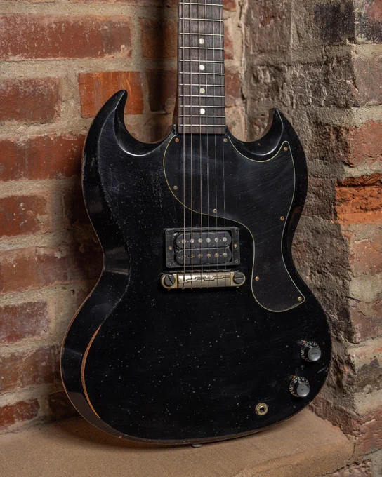 A black Gibson SG electric guitar leaning against a brick wall. The guitar has a worn finish, dual cutaways, and two control knobs.