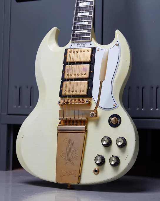 A white Gibson SG electric guitar with a vintage design, featuring gold hardware and a white pickguard. The guitar has a distinct body shape and multiple control knobs. It rests vertically against a dark background.