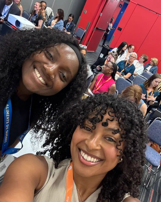 Two women with curly hair take a selfie in a conference room filled with seated attendees engaged in conversation.