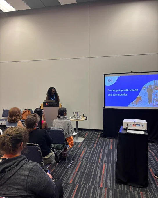 Speaker presenting on co-designing with schools and communities to an attentive audience in a conference room.