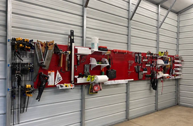 A well-organized workshop featuring red Wall Control metal pegboards mounted on a metal garage wall, holding clamps, saws, hand tools, power tools, shelves, and storage accessories in a clean and efficient layout.