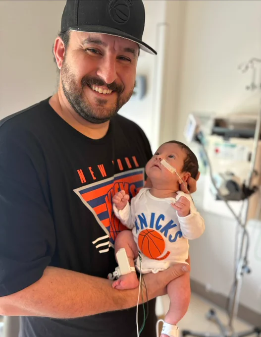 A smiling bearded man in a black hat and a 'NEW YORK' shirt holds a baby. The baby, wearing a white 'KNICKS' onesie, looks up and has a nasal cannula and monitoring wires on its legs. They are in what appears to be a medical setting.