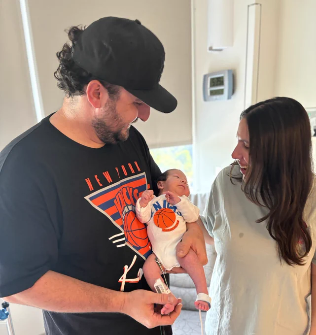 A man in a black New York Knicks shirt and cap holds a newborn baby with medical sensors on its feet. The baby wears a white onesie with \"KNICKS\" and \"1ST GAME WITH DAD 6.X.22\". A woman with long dark hair smiles at them. A wall clock shows 5:35.