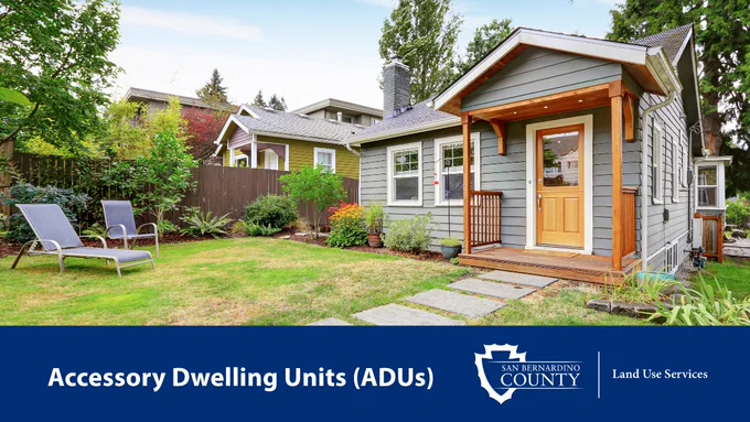 A gray house with a wooden porch is surrounded by a neatly maintained yard with two lounge chairs. A sign reads "San Bernardino County Land Use Services, Accessory Dwelling Units (ADUs)."