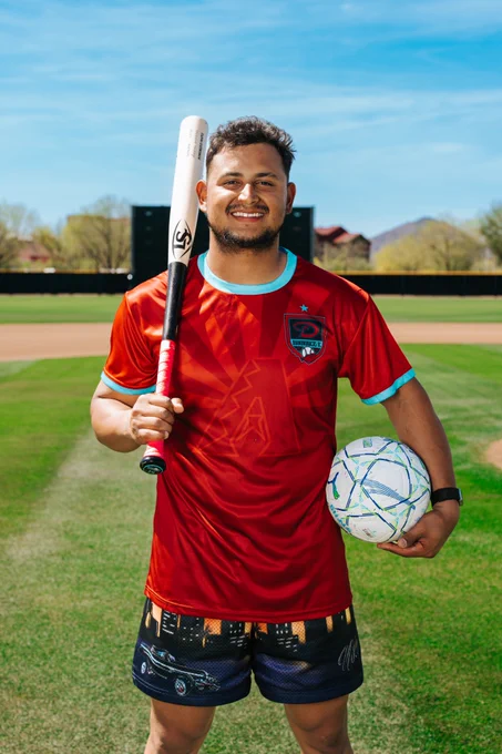 Gabriel Moreno smiles at the camera while resting a bat on his shoulder and a soccer ball on his hip. He is wearing a red and teal Gabriel Moreno Soccer Jersey. 