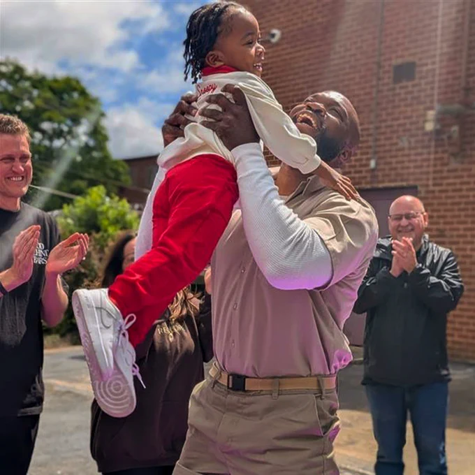 The image shows a joyful person lifting a child wearing a white shirt and red pants outside a brick building. People around them are clapping and smiling, enjoying the moment. It appears to be a sunny day.