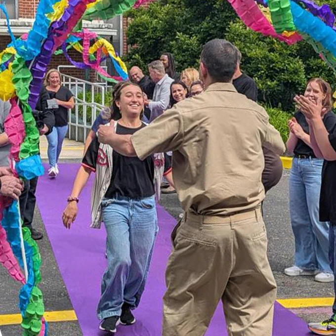 A person walks on a purple carpet surrounded by colorful, twisted paper streamers, with others clapping and smiling around them. The scene is lively and celebratory, taking place outdoors.