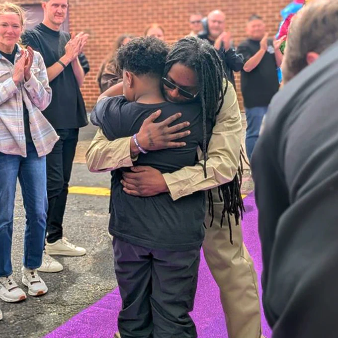 A person with long hair wearing sunglasses hugs a child on a purple carpet. People around them are clapping.