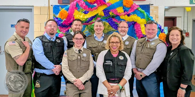 A group of people wearing uniform vests stand together smiling in front of a vibrant, colorful background decorated with paper flowers. 