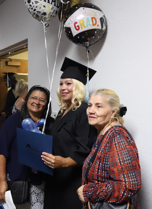 Graduate poses with two others while holding celebratory balloons and diploma.