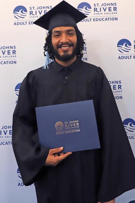 Graduate in cap and gown holding a St. Johns River State College Adult Education diploma folder against a branded backdrop.