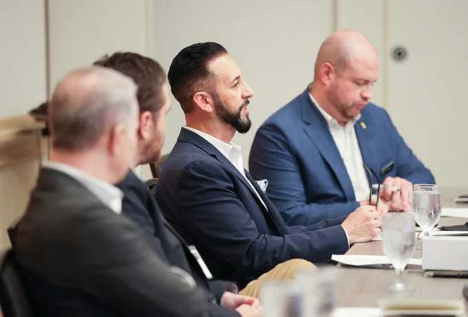 A man in a navy blazer speaks during a conference discussion while seated at a meeting table with other attendees listening nearby.