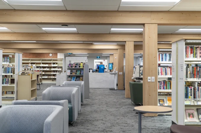 A spacious library interior with wooden beams and several rows of bookshelves filled with books. Comfortable seating areas with gray armchairs are visible, and a library desk can be seen in the background.