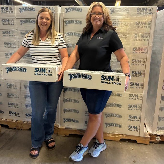 Two people stand smiling in a warehouse, each holding a box labeled "Sun Meals-To-Go" and "Power Packs." There are stacks of similar boxes in the background.