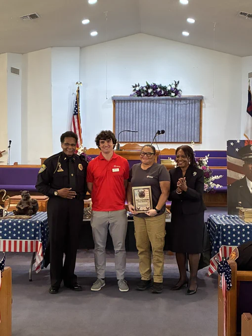 A group of four individuals stands together indoors with a table displaying patriotic decorations nearby. One person is holding a framed item. There is a backdrop with a U.S. flag and a large portrait in the background. The setting appears to be a formal event.