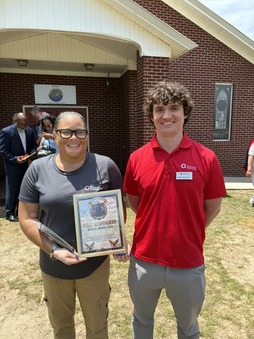 Two people are standing outside a brick building. One is holding a framed certificate titled "First Responders Award." Both are smiling. Other individuals are visible in the background.