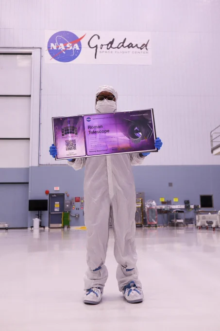 A person holds a large, purple "boarding pass" for NASA's Nancy Grace Roman Space Telescope in Goddard's large cleanroom wearing a full white cleanroom suit. The boarding pass has the detector logo for Roman, an artist concept image of Roman, as well as a QR code, the telescope, and the NASA logo. "Roman Telescope" is written where the name would go.