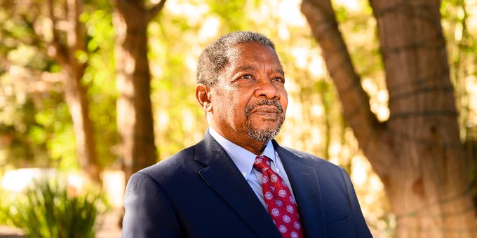 Man in a navy blue suit and red patterned tie standing outdoors with trees and sunlight in the background.