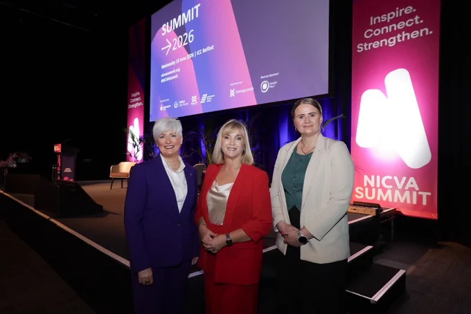 Image of three people - left to right Junior Minister Joanne Bunting, Celine McStravick CEO of NICVA and Junior Minister Aisling Reilly standing in front of  a stage with screen in background