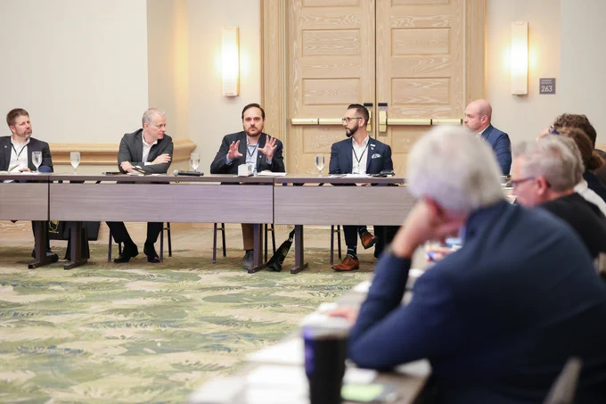 Participants sit around a U-shaped conference table as a panelist gestures while speaking during a group discussion in a meeting room.