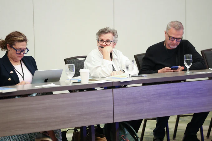 Three conference attendees sit at a meeting table; one works on a laptop, one listens thoughtfully with a pen in hand, and another looks at a smartphone.