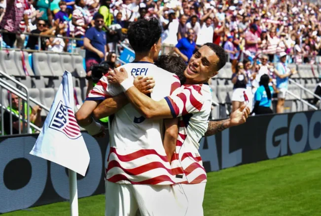 A group of soccer players in striped jerseys celebrate a goal by hugging near the corner flag during a sunny match. The crowd cheers in the background, adding to the festive atmosphere.
