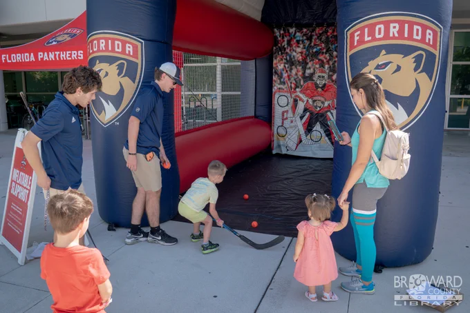 A young boy using a hockey stick to smack a ball at an inflatable goalie while his family watches