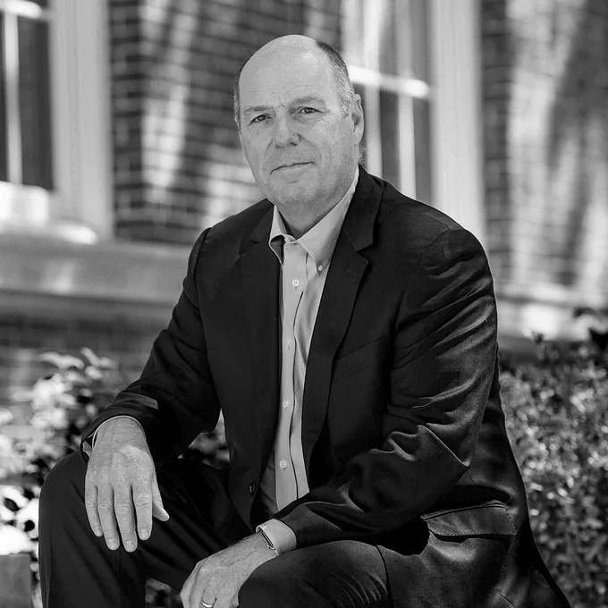 A black and white portrait of Martin A. Schmidt, president of RPI. He is wearing a dark suit and light button-down shirt, sitting outdoors with a brick building and foliage blurred in the background.