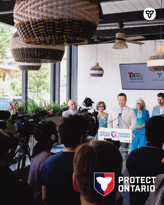 Minister Stan Cho stands at a podium during a press event for Taste of the Danforth, with media cameras in the foreground and attendees behind. Event branding appears on a screen, and the Protect Ontario logo is on the podium and in the corner.