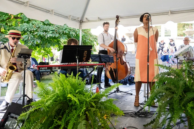 Jazz band performing under a white tent with a female vocalist in an orange dress and musicians playing saxophone, keyboard, upright bass, and drums.