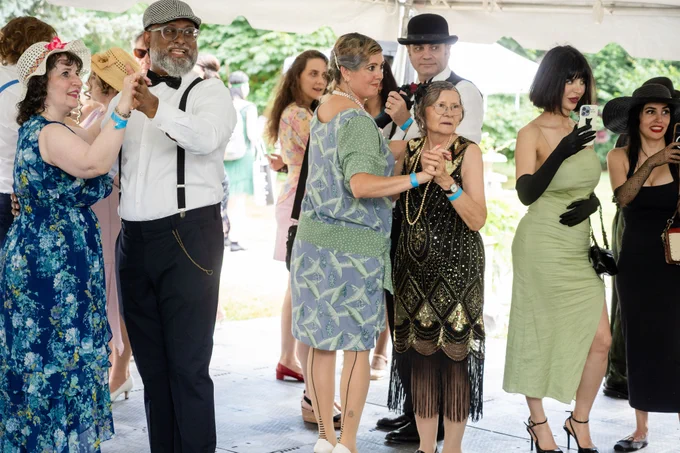 Group of people dressed in vintage 1920s attire dancing and socializing under a white tent at an outdoor event.