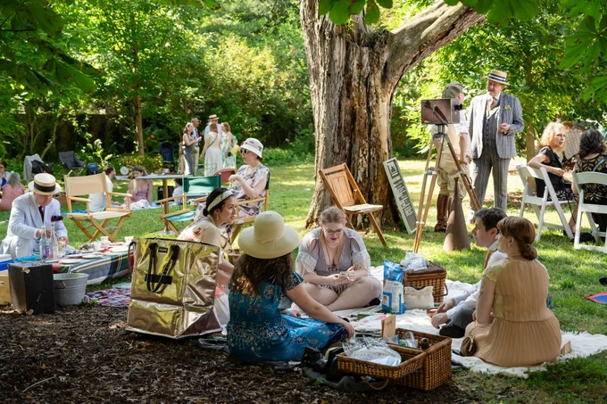 Group of people enjoying a sunny picnic under large trees with blankets, baskets, and vintage decor in a lush garden setting.