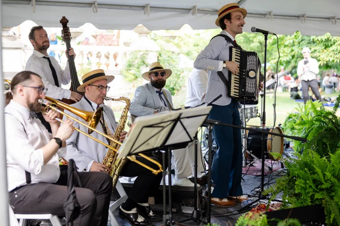 A jazz band performs under a white tent with musicians playing saxophone, trumpet, double bass, and accordion on a sunny day.