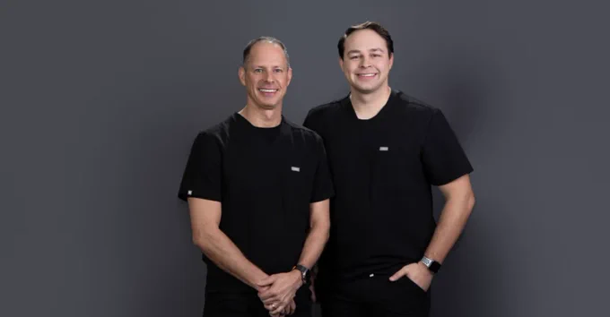 Two smiling men in black short-sleeved shirts with small chest logos stand against a dark gray background. The man on the left has grey hair & clasped hands; the man on the right has dark hair, a hand in his pocket & a smartwatch.
