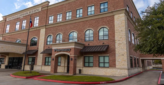 The Magnolia Landmark Building, a multi-story structure with red brick and light stone accents. It features various windows, some arched. A Texas flag flies on a pole in front. The building also has a 'Pharmacy' sign. Green landscaping and a paved area with a red curb are visible.