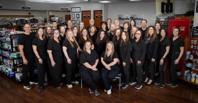 A large group of about 30 adults, mostly women, wearing black scrubs, smiling in a brightly lit pharmacy. Visible signs include \"Magnolia Pharmacy,\" \"Drop Off,\" \"Pick Up,\" and \"Consultation.\" Pharmacy shelves with various products are in the background.