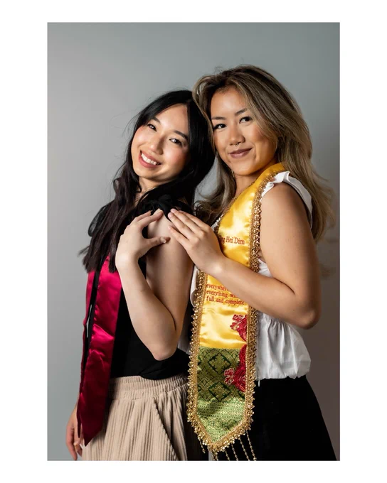 Two people stand closely together, smiling. Both wear decorative stoles. The background is plain.