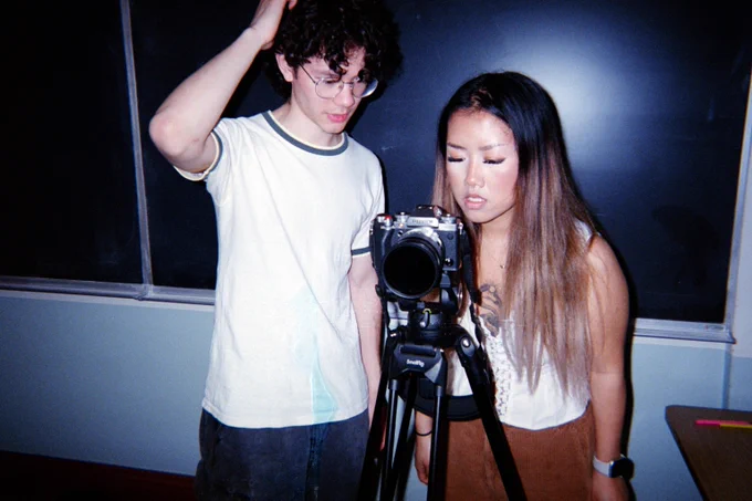 Two people stand in front of a camera on a tripod. One person is touching their head, while the other looks closely at the camera. They are in a room with a chalkboard.