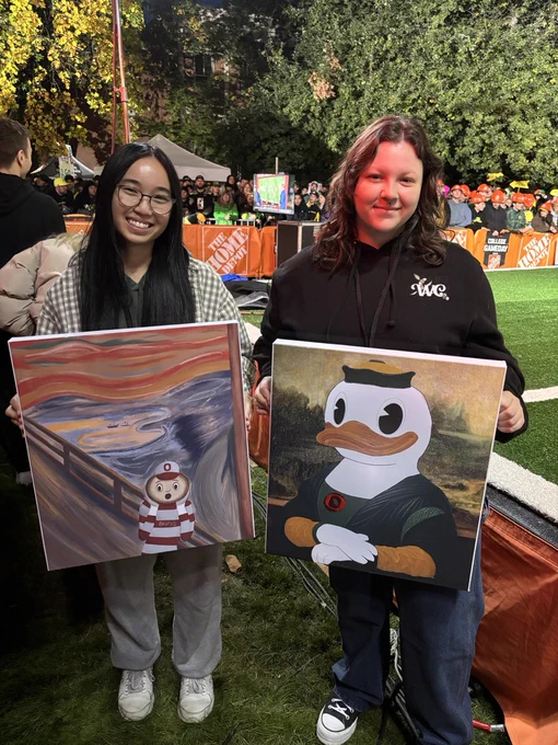 Two people are standing outdoors, each holding a painting. The person on the left holds a painting resembling Edvard Munch's "The Scream" with a small striped figure in the foreground. The person on the right holds a painting of The Duck mascot as the Mona Lisa. A crowd and some trees are visible in the background.