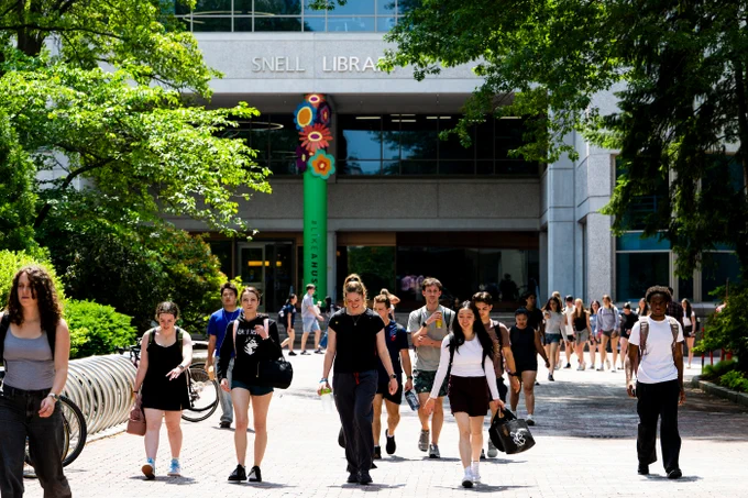 Diverse young adults walk on a sunny brick path in front of Snell Library. Green trees frame the scene. A green column with colorful flowers and '#LIKEAHUSKY' stands near the entrance.