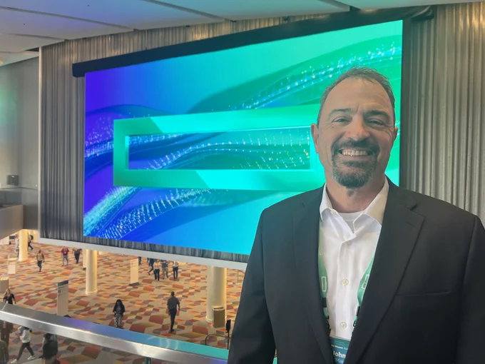 John McMains standing in front of a large digital screen displaying a blue and green abstract design at HPEDiscover venue.