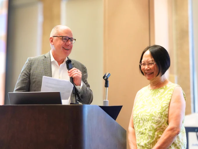 Man at podium speaking and smiling, woman next to him smiling