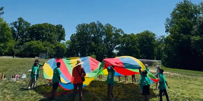 Children playing with a large colorful parachute on a sunny grassy field surrounded by trees.