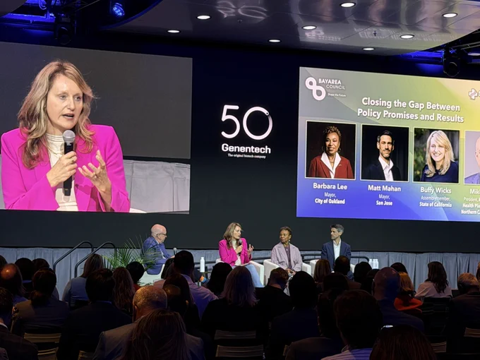 Assemblymember Wicks speaks on stage at an event hosted by Bay Area Council. She is seated on a moderated panel alongside Mayor Barbara Lee and Mayor Matt Mahan, with a large screen behind the stage projecting her image to the audience.