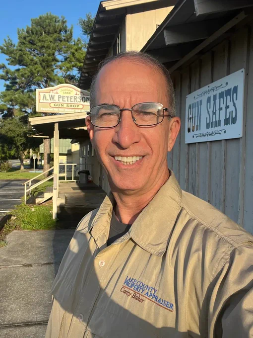 A man in glasses and a tan shirt smiling while standing outside a gun shop during the daytime. The shop sign is visible in the background.