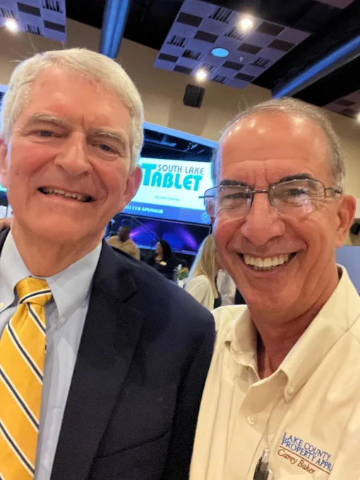 Two men are taking a selfie at an event with a banner displaying 'South Lake Tablet' in the background. The atmosphere is professional and cordial.